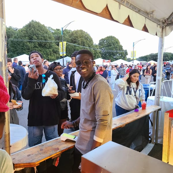 A smiling man in glasses stands behind a counter at an outdoor food festival with many people.