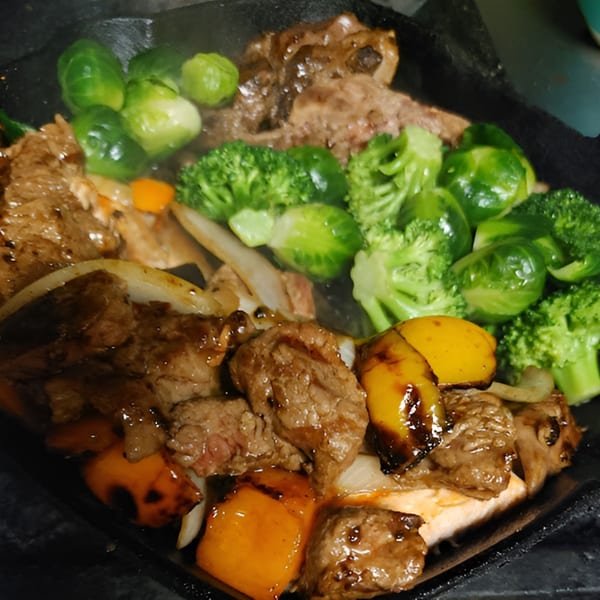 Sizzling beef and vegetables, including broccoli, Brussels sprouts, and bell peppers, on a dark plate.