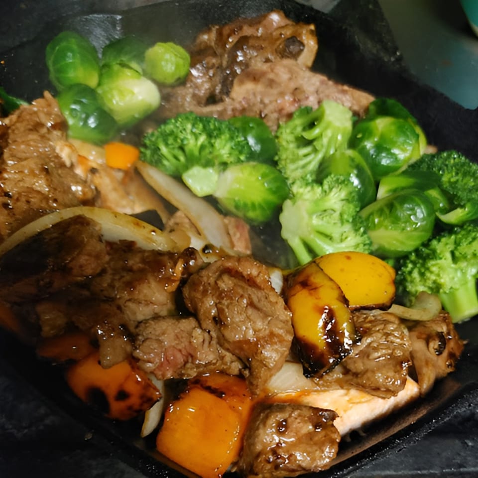 Sizzling beef and vegetables, including broccoli, Brussels sprouts, and bell peppers, on a dark plate.