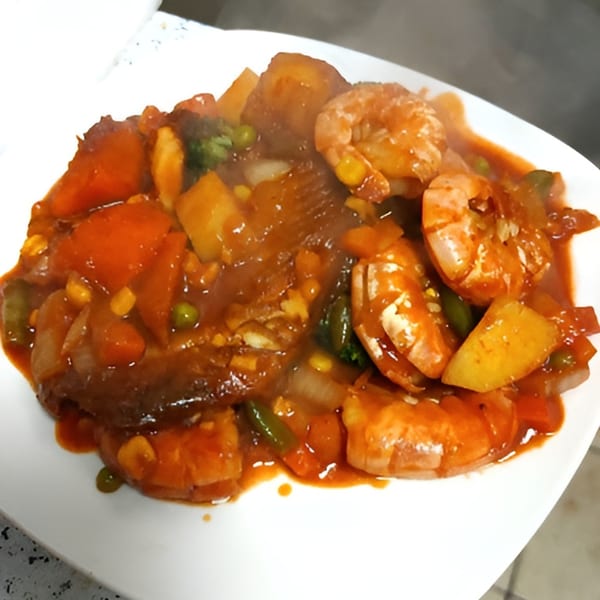 A steaming plate of Badou Senegalese Cuisine's Thieboudienne, featuring fish, shrimp, and vegetables in a rich red sauce.