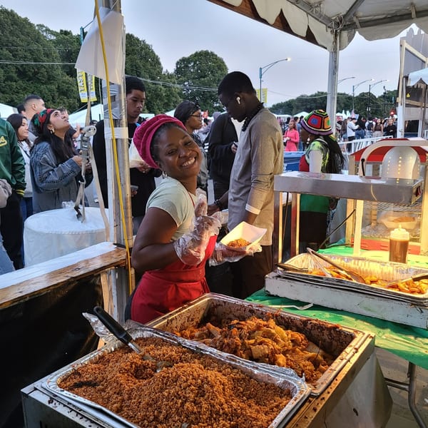 A smiling woman in a red apron serves Senegalese food from large trays at an outdoor event.