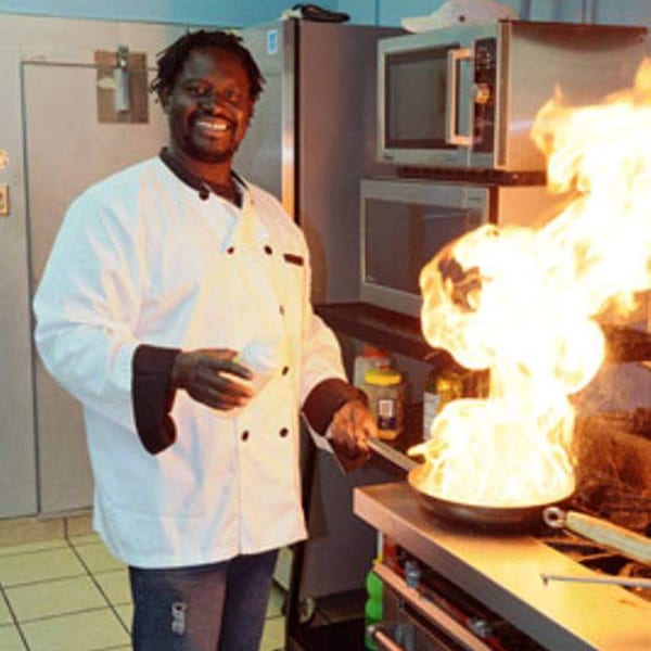 A smiling chef in a white coat flambés food in a pan with a large flame in a kitchen.